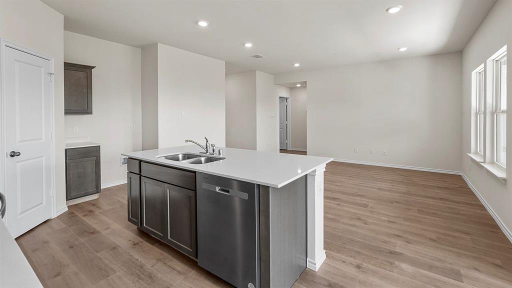 Kitchen featuring a central island with a solid-surface countertop, dual-basin stainless steel sink, built-in dishwasher, shaker-style cabinetry, and wood-finish flooring