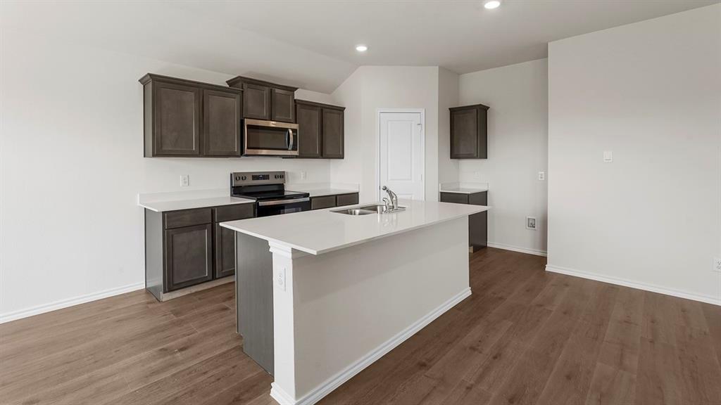 Kitchen featuring dark-finish cabinetry, light-toned countertops, a center island with an integrated dual-basin sink, stainless steel appliances, and wood-finish flooring