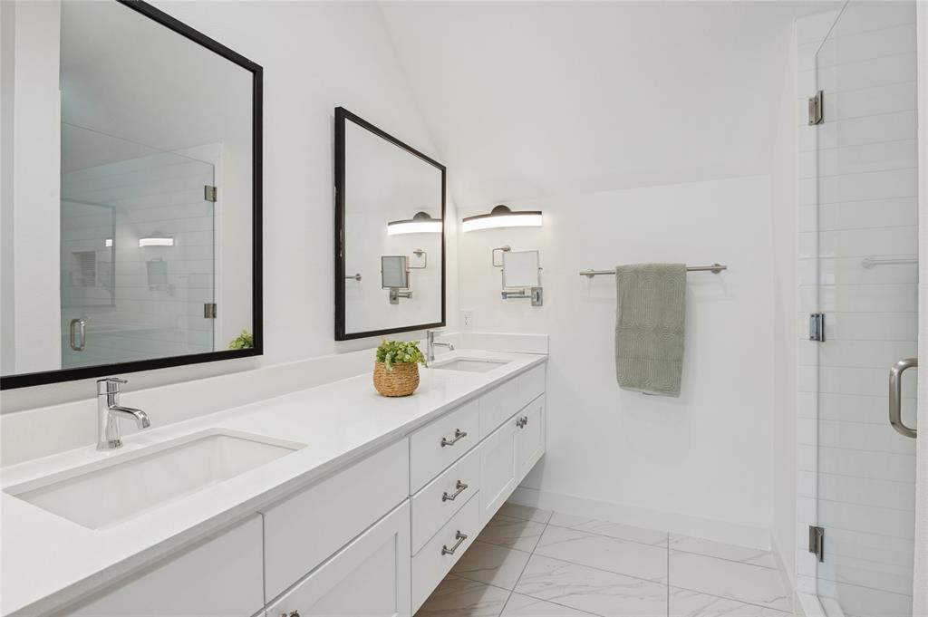 Dual-sink vanity with a solid-surface countertop, white cabinetry with metal hardware, framed mirrors, wall-mounted lighting, and a glass-enclosed shower with tiled flooring