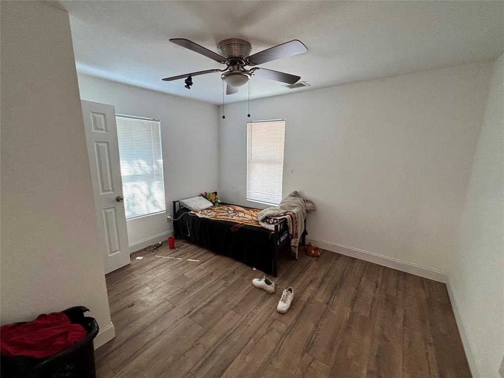 Neutral interior room featuring wood-finish flooring, two windows with blinds, a multi-panel interior door, and a ceiling fan with integrated light fixture