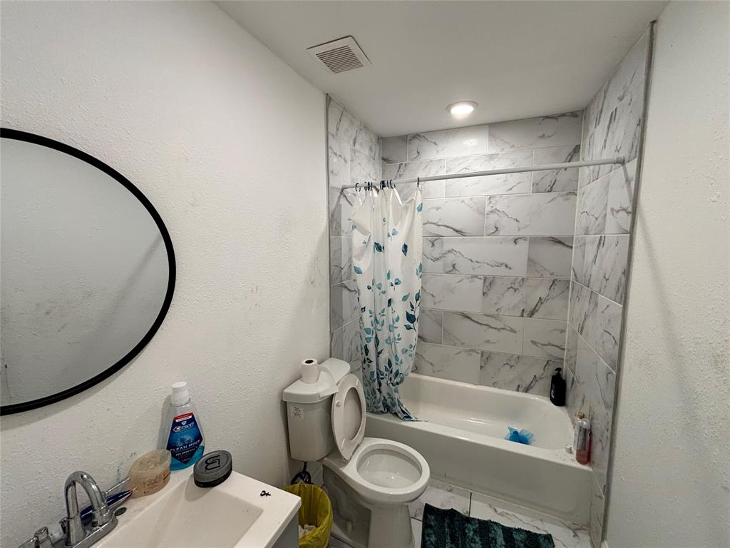 Full bathroom featuring a shower-tub combination with large-format stone-patterned wall tile, a round wall-mounted mirror, and a vanity with a chrome faucet