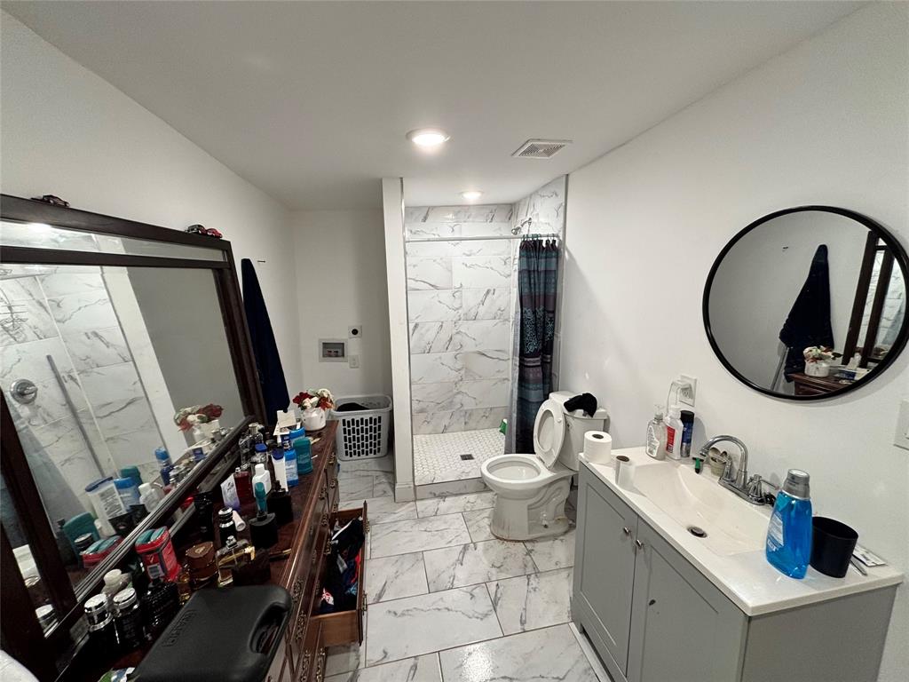 Bathroom featuring a gray vanity with a white countertop, an integrated sink with a chrome faucet, large-format tile flooring, a walk-in shower with tiled walls, and a circular wall-mounted mirror