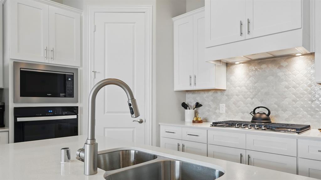 White cabinetry, stone-finish countertops, stainless steel dual-basin sink, scalloped tile backsplash, and built-in stainless steel double wall oven and microwave