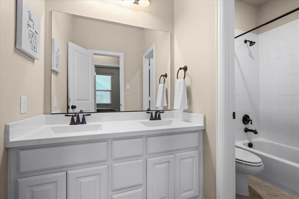 Dual-sink vanity with white cabinetry, solid-surface countertop, bronze fixtures, large vanity mirror, and a tub-shower combination featuring white tile surround