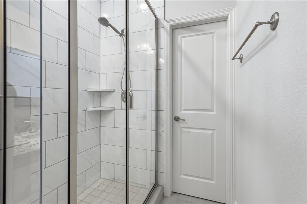 Walk-in shower featuring a glass enclosure, wall-mounted showerhead, built-in corner shelving, and tile-surround walls, complemented by a solid-core paneled door and metal wall-mounted towel bar