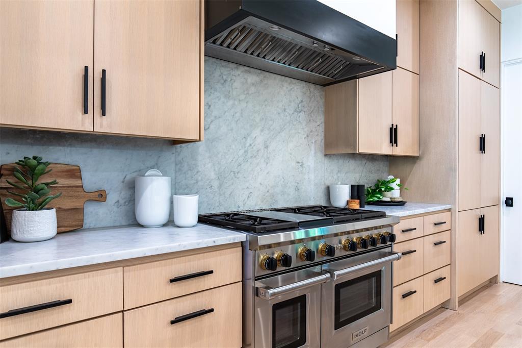 Flat-panel light wood cabinetry with matte black hardware, stone slab backsplash, stone countertops, professional-grade stainless steel range with dual ovens, and a stainless steel range hood