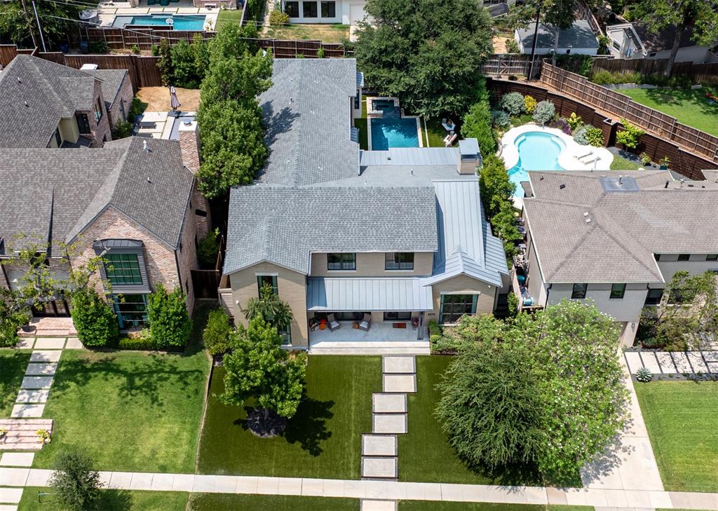 Aerial perspective of a multi-story residence featuring a gray shingle and metal roof, a rear patio with stone-finish pavers, a built-in swimming pool, and manicured landscaping with mature trees