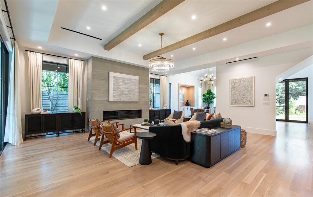 Spacious living area featuring light-toned wood-finish flooring, a coffered ceiling with exposed wooden beams, recessed lighting, a linear fireplace with a textured stone surround, and floor-to-ceiling iron-framed glass doors