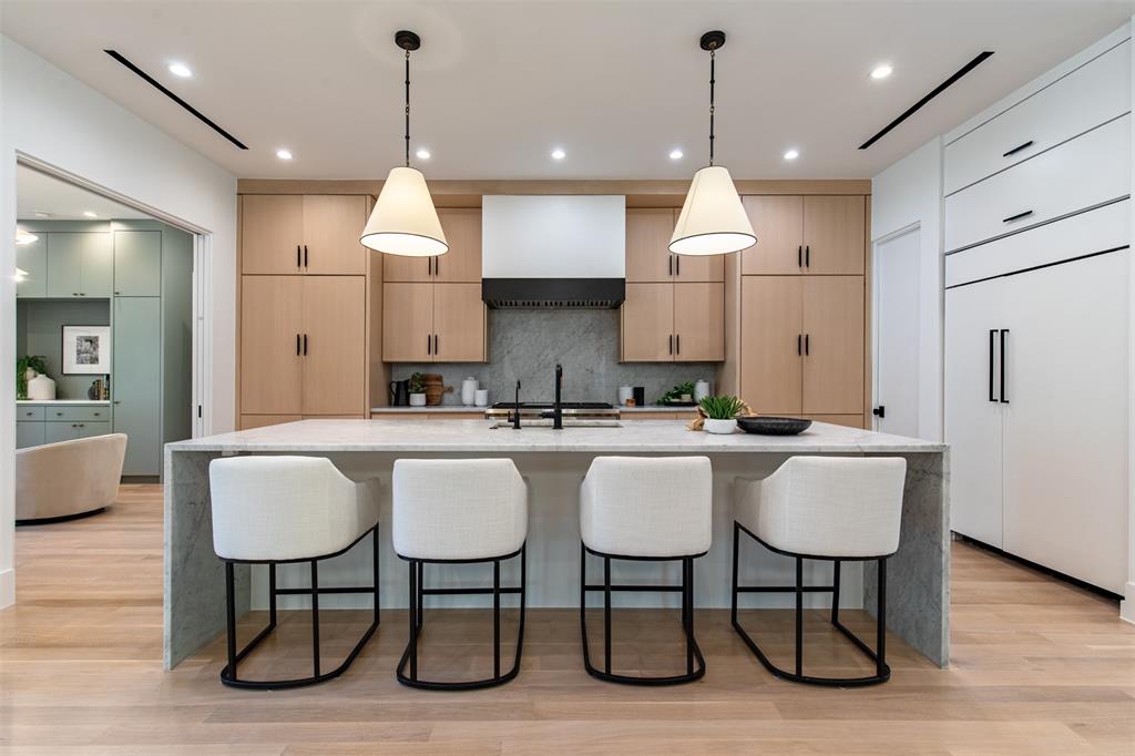 Kitchen featuring custom light wood cabinetry, a central island with a waterfall-edge countertop, dual pendant lights, a stone backsplash, and recessed ceiling lighting