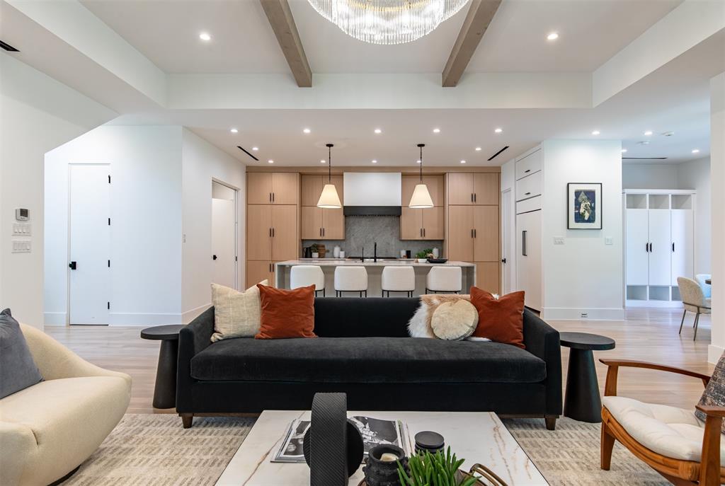 Open-concept kitchen and living area featuring tray ceilings with wooden beams, custom cabinetry, a central island with stone surfacing, and a crystal chandelier