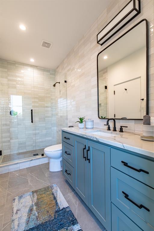 Bathroom featuring a blue-toned vanity with matte black hardware, stone-patterned countertop with an integrated rectangular sink, walk-in shower with a glass enclosure, and wall-to-wall light-toned tile backsplash