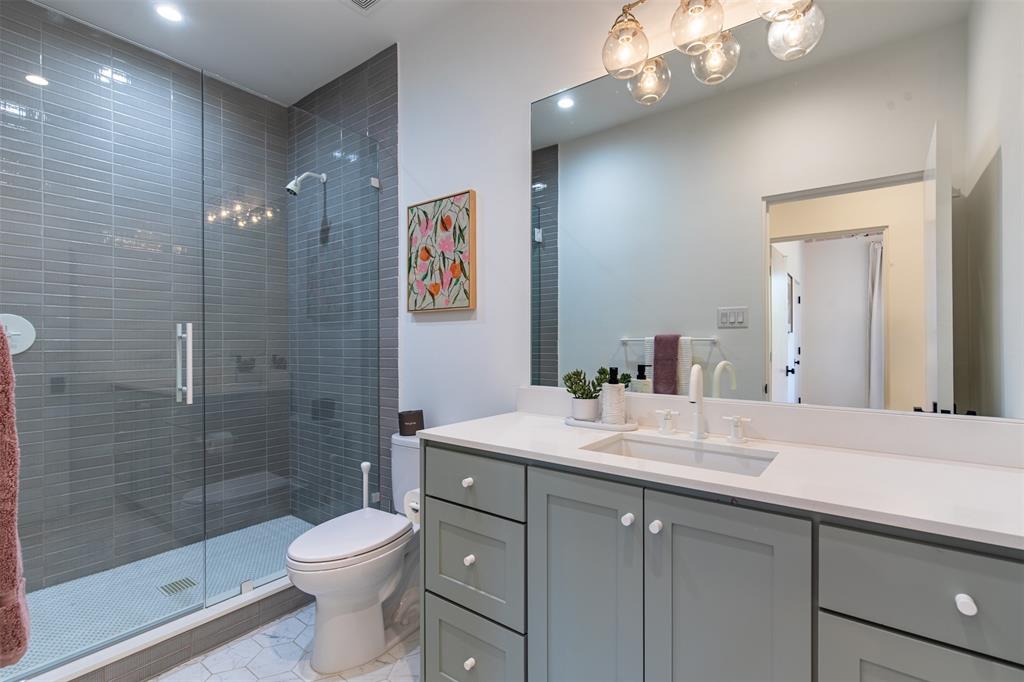 Bathroom featuring a grey shaker-style vanity with a solid surface countertop, an undermount sink, a walk-in glass-enclosed shower with horizontal wall tile, and hexagonal floor tile