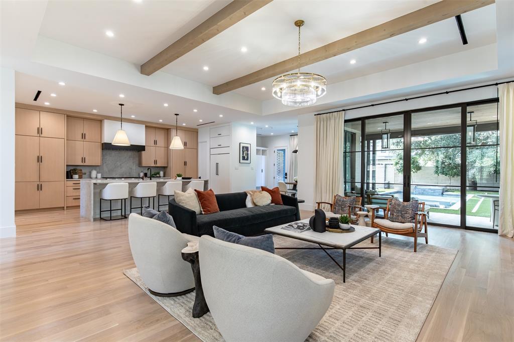 Open-concept living space featuring wood-finish flooring, a coffered ceiling with exposed wood beams, a custom kitchen with light-tone cabinetry, a central stone-top island, and floor-to-ceiling metal-framed glass doors