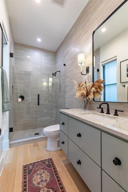 Custom vanity with stone countertop and matte black fixtures, walk-in shower with frameless glass enclosure, tiled wall accents, and wood-finish flooring
