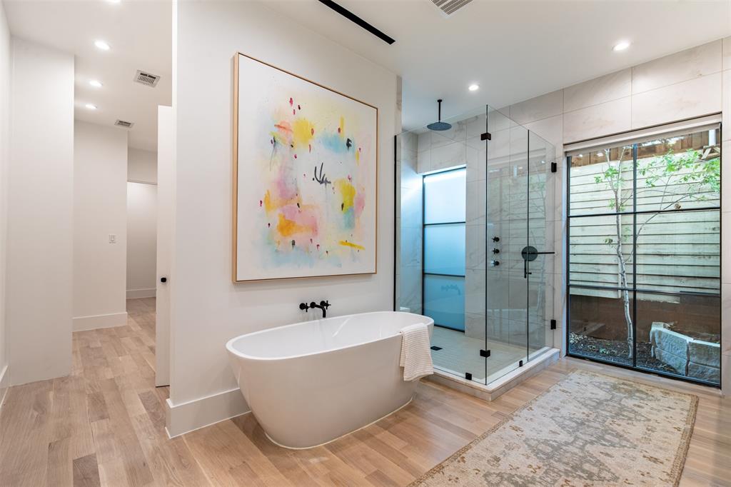 Freestanding soaking tub with matte black wall-mounted faucet, glass-enclosed walk-in shower with rainfall showerhead, large-format tile surround, and wood-finish flooring