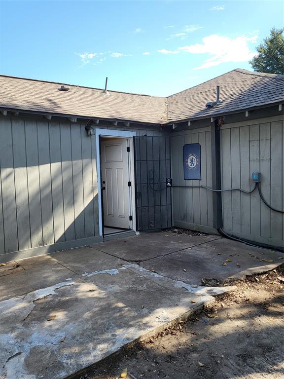 Exterior concrete patio featuring vertical wood-style siding, an asphalt shingle roof, and a metal security gate