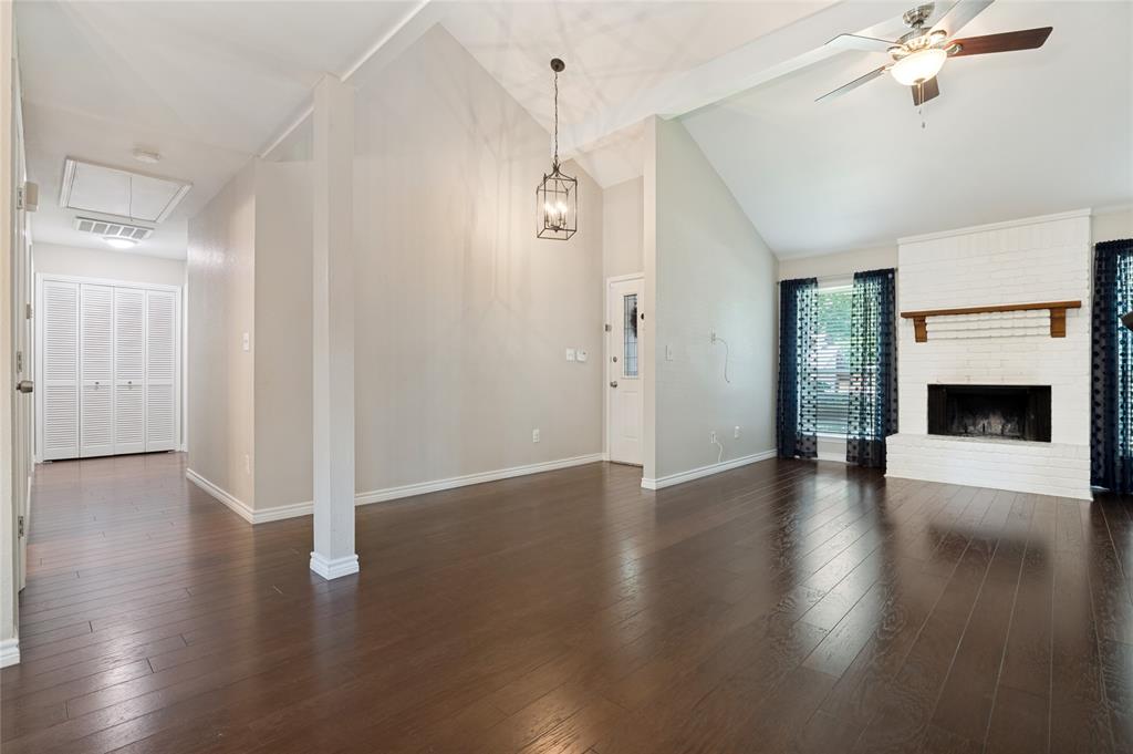 Vaulted ceiling with exposed beam, wood-finish flooring, white brick fireplace with wood mantle, ceiling fan with integrated lighting, and white bi-fold closet doors