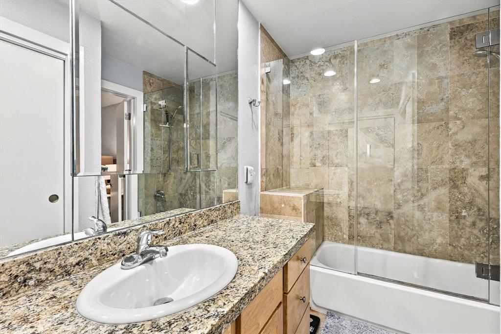 Bathroom featuring a vanity with stone countertop, an integrated oval sink, full-width mirror, and a tub-shower combination enclosed by a glass panel with floor-to-ceiling stone-tile surround