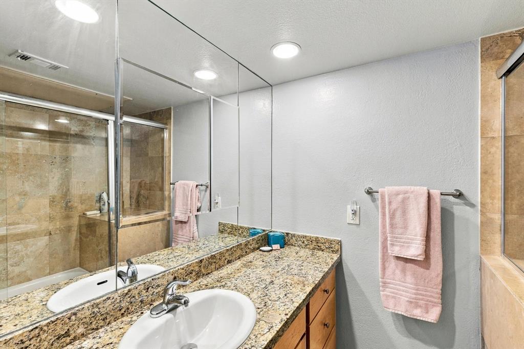 Full-width wall mirror, stone-surfaced vanity top with an integrated oval sink, wood-finish cabinetry, and a walk-in shower enclosure featuring tiled walls and sliding glass doors