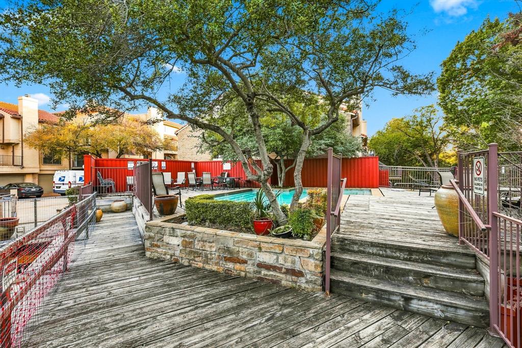 Outdoor pool area featuring a stone-encased planter, expansive wood decking, metal perimeter fencing, and mature canopy trees