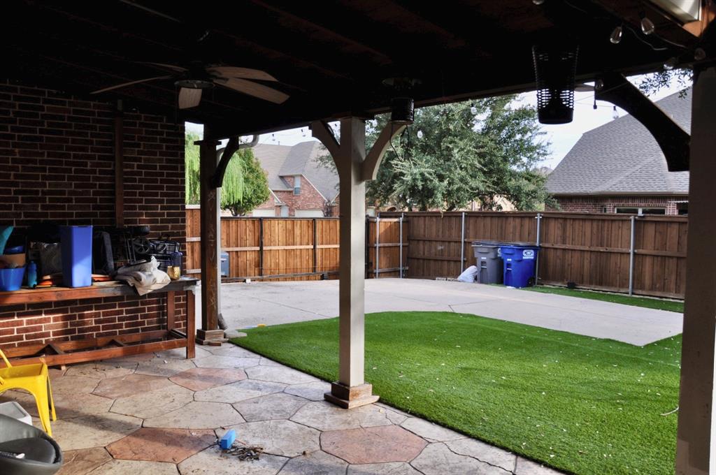 Covered patio featuring stamped concrete flooring, a ceiling fan, and exposed brick walls, overlooking a backyard with synthetic turf and a wood privacy fence