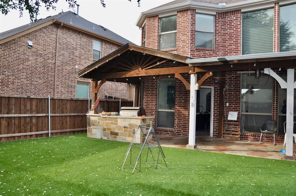 Backyard featuring a covered patio with timber framing, stone-clad outdoor kitchen, brick exterior, and synthetic turf lawn
