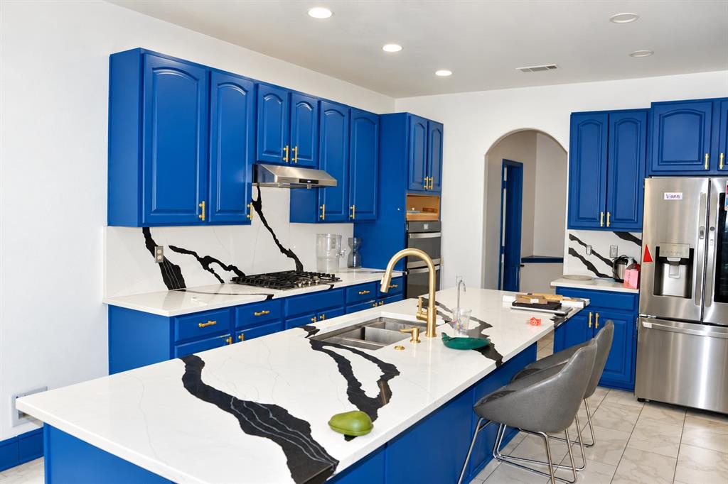 Kitchen featuring bold blue cabinetry, veined stone-finish countertops with matching backsplash, stainless steel appliances, a center island with an integrated dual-basin sink, and brass-finish hardware