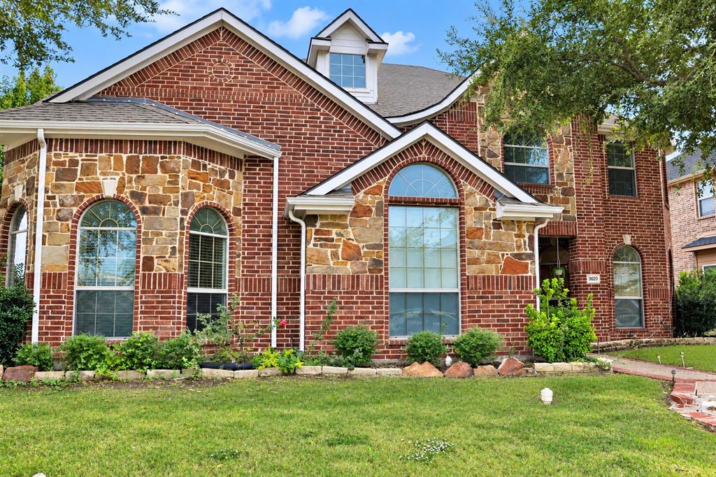 Two-story exterior featuring red brick and natural stone masonry, arched window detailing, a steep gabled roofline, and a manicured front lawn