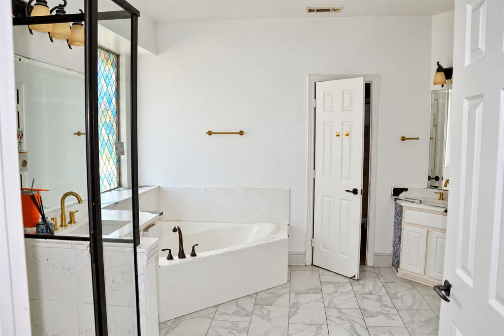 Soaking tub with diamond-pattern tile surround, vanity featuring a rectangular undermount sink, multi-tone stained glass window, brass-finish hardware, and stone-finish flooring