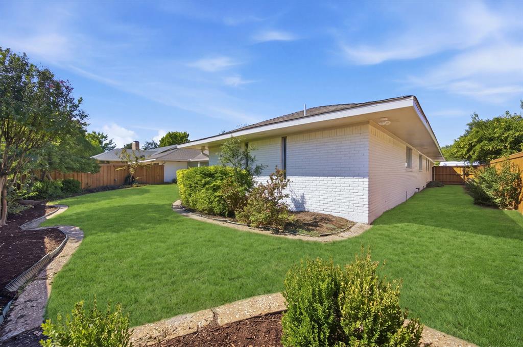White brick exterior featuring a hip roof, expansive lawn, stone-border garden beds, and wood privacy fencing