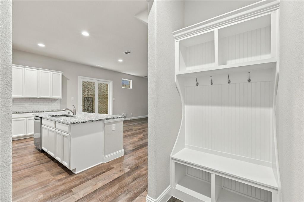 Built-in white mudroom bench featuring beadboard detailing, upper storage cubbies, and garment hooks, complemented by an open-concept kitchen area with white cabinetry, stone-finish countertops, and wood-finish flooring