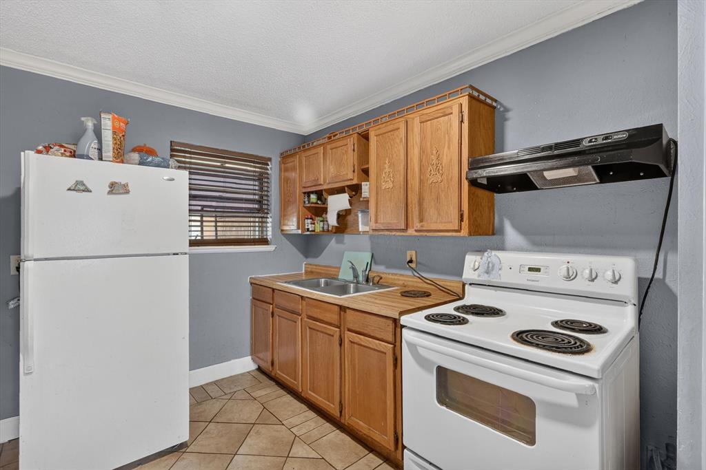 Kitchen featuring natural wood cabinetry, dual-basin stainless steel sink, tile flooring, and wall-mounted range hood
