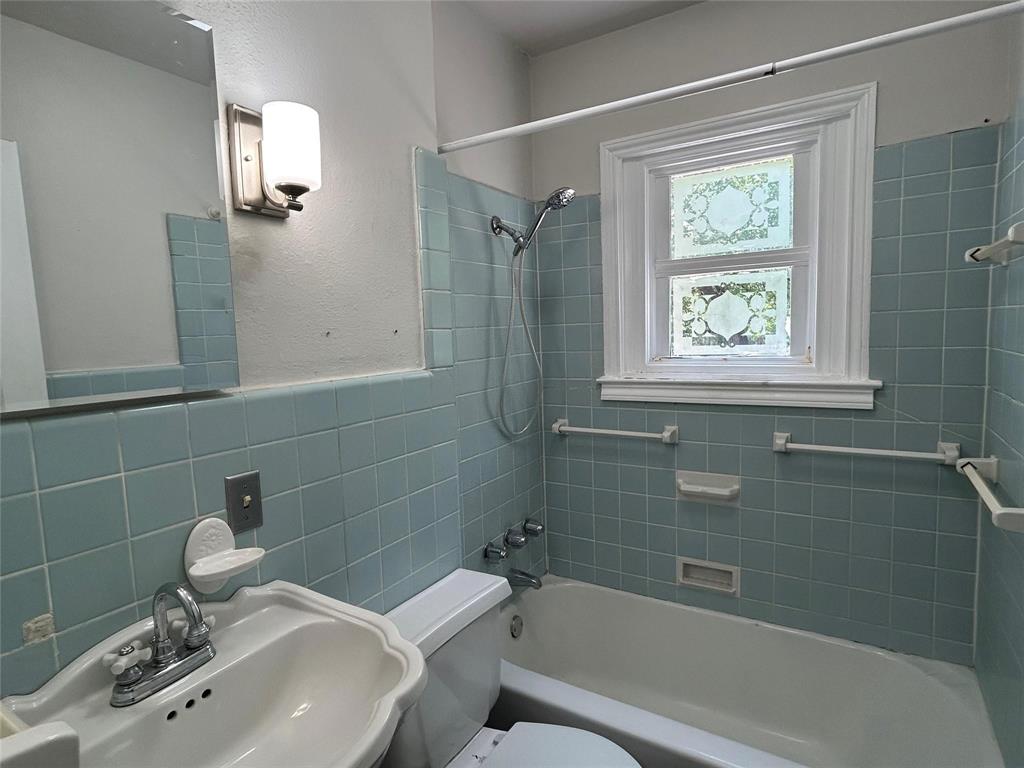 Bathroom featuring blue tile wall surround, integrated bathtub and shower combination, wall-mounted light fixture, pedestal sink, and privacy window with decorative glass detailing