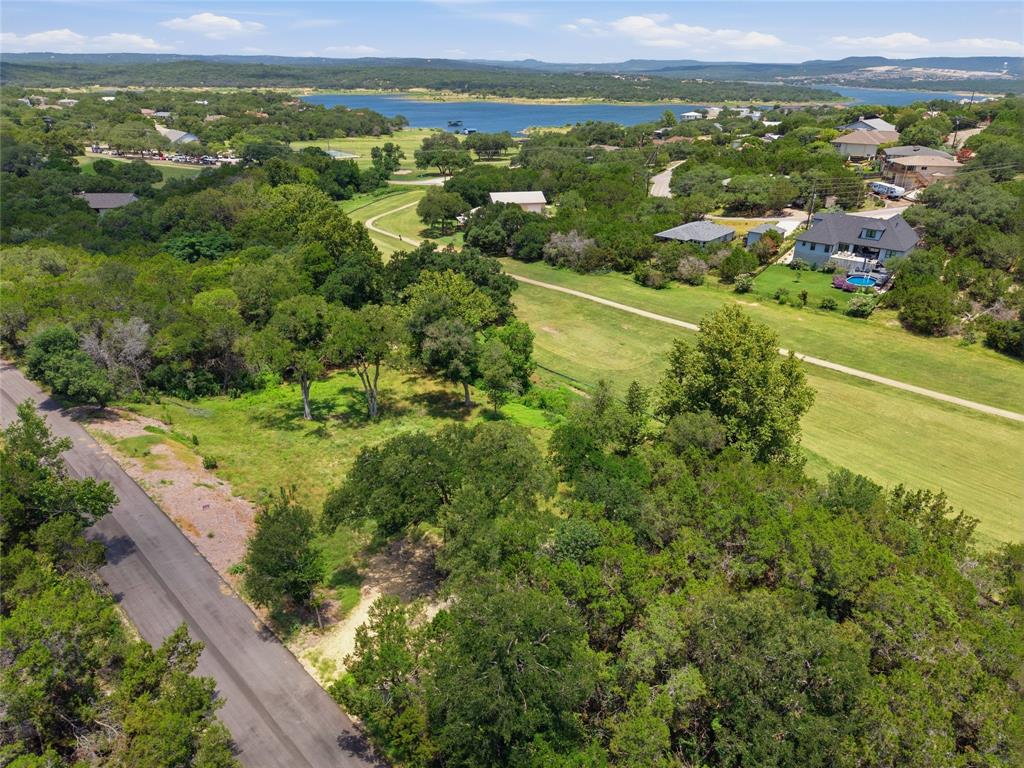 Aerial perspective of a sloping lot featuring mature native trees, paved street access, and a backdrop of expansive water scenery