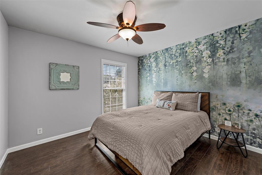 Room featuring wood-finish flooring, a multi-blade ceiling fan with dual-light fixtures, a single-hung window with horizontal blinds, and a botanical-patterned accent wall
