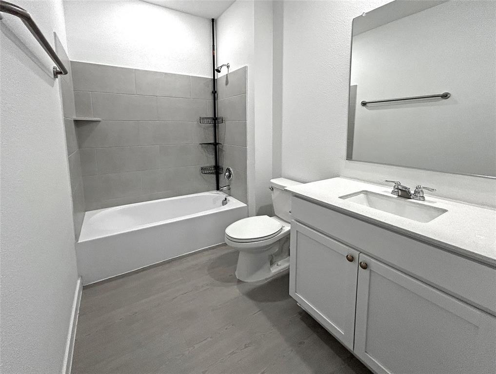 A full bathroom features a white vanity with a light-toned countertop, integrated rectangular sink, large wall-mounted mirror, luxury vinyl plank flooring, and a bathtub with gray tile surround