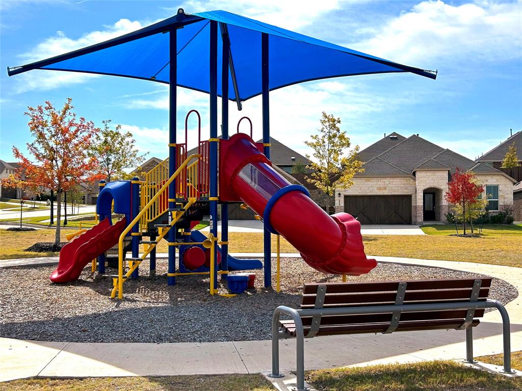 Front of house with a stone exterior as seen from the community recreational area. The recreation area features a structured play system with a blue shade canopy, integrated slides, a gravel base, a fixed metal and wood bench and interconnected sidewalks.