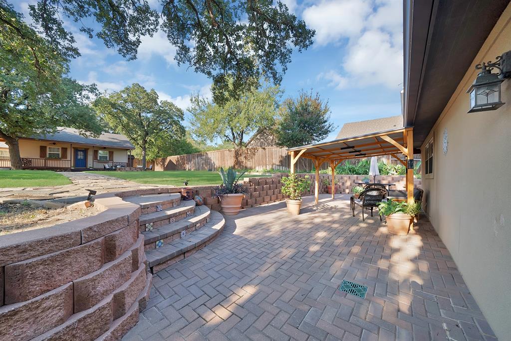 Herringbone-patterned paver patio featuring a timber-framed pergola, segmental retaining walls, decorative tiled steps, and a sodded lawn
