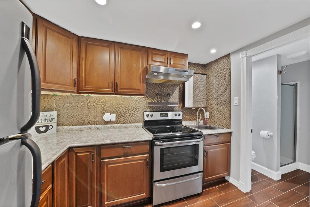 Kitchen featuring stained wood cabinetry, stone-finish countertops, textured tile backsplash, stainless steel range with overhead vent hood, and wood-finish flooring