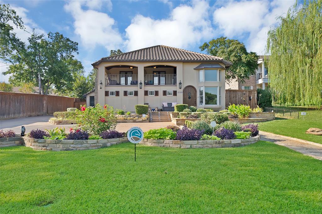 Stucco exterior featuring a tiled roof, upper-level balcony with metal railing, arched entryway, and tiered stone retaining walls with landscape beds