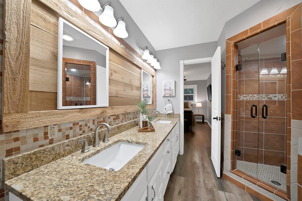Dual-sink vanity with stone countertop, wood-plank wall accent, glass-enclosed shower with tiled surround, and wood-finish flooring