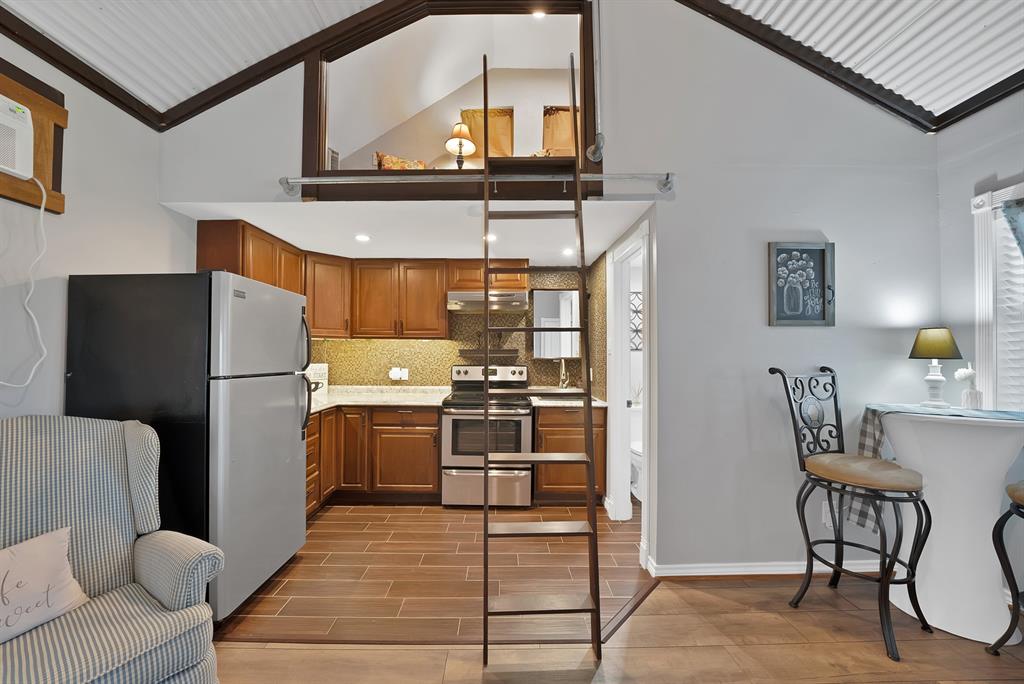 Kitchen featuring wood cabinetry, stainless steel appliances, tile backsplash, and wood-finish flooring, complemented by a structural ladder providing access to an elevated mezzanine level under a vaulted ceiling