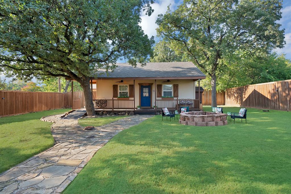 Exterior facade featuring a covered front porch, stone-paved walkway, fire pit, mature trees, and wood-panel privacy fencing