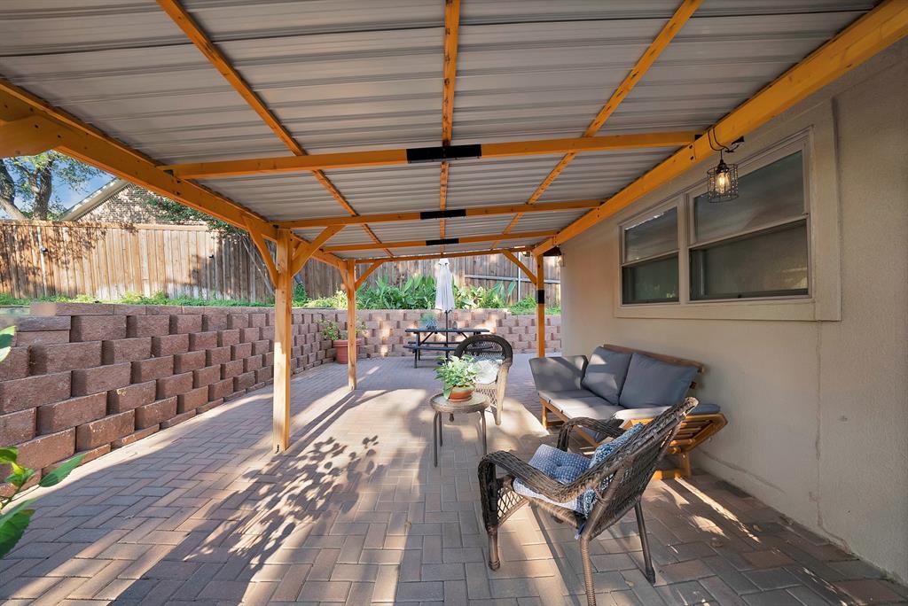 Covered outdoor patio featuring a metal roof, wood-frame support beams, interlocking paver flooring, and a tiered stone retaining wall
