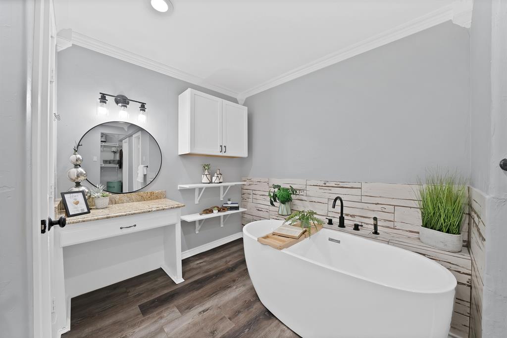 Freestanding soaking tub featuring a matte black deck-mounted faucet, wood-finish flooring, white vanity with natural stone countertop, circular wall mirror, and crown molding