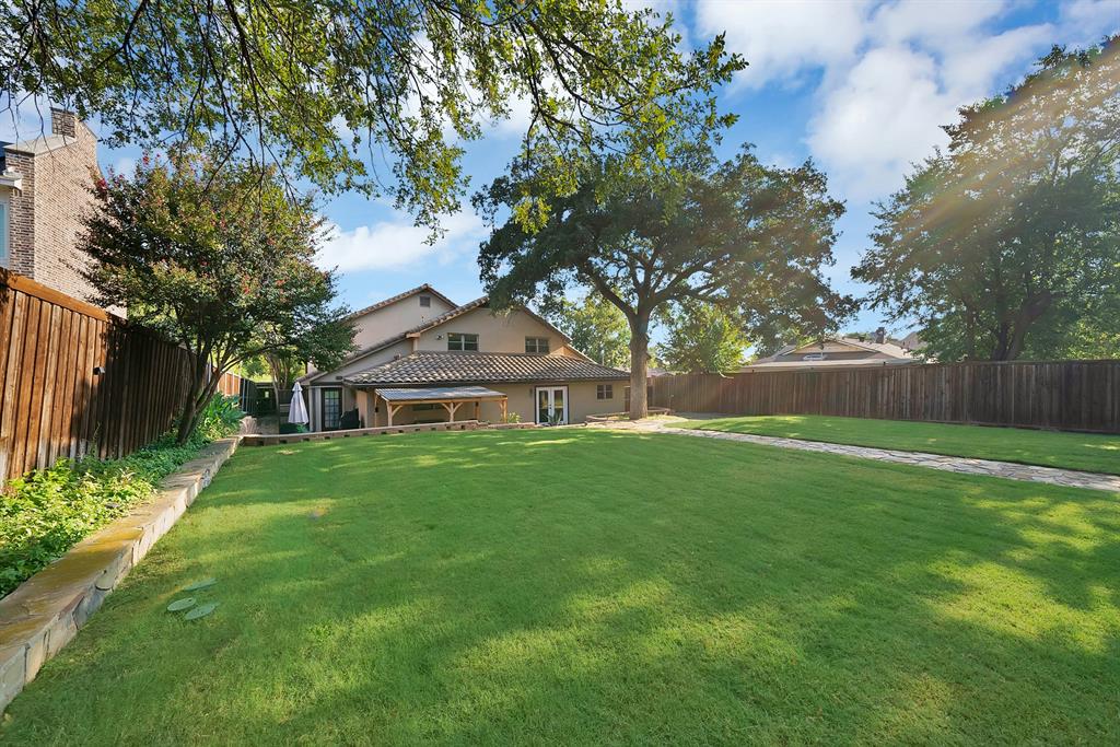 Expansive backyard featuring a level lawn, stone retaining walls, stone-paved walkway, wood privacy fencing, and mature canopy trees