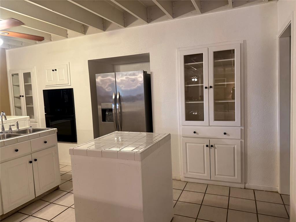 Kitchen featuring white cabinetry with glass-front display uppers, tile countertops, stainless steel appliances, ceramic tile flooring, and exposed ceiling beams