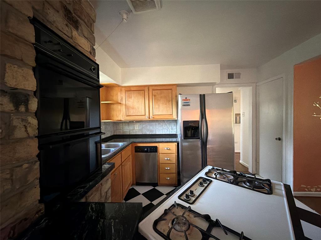 Kitchen featuring a stone-faced wall, natural wood cabinetry, dark stone countertops, stainless steel appliances, and a built-in cooktop island