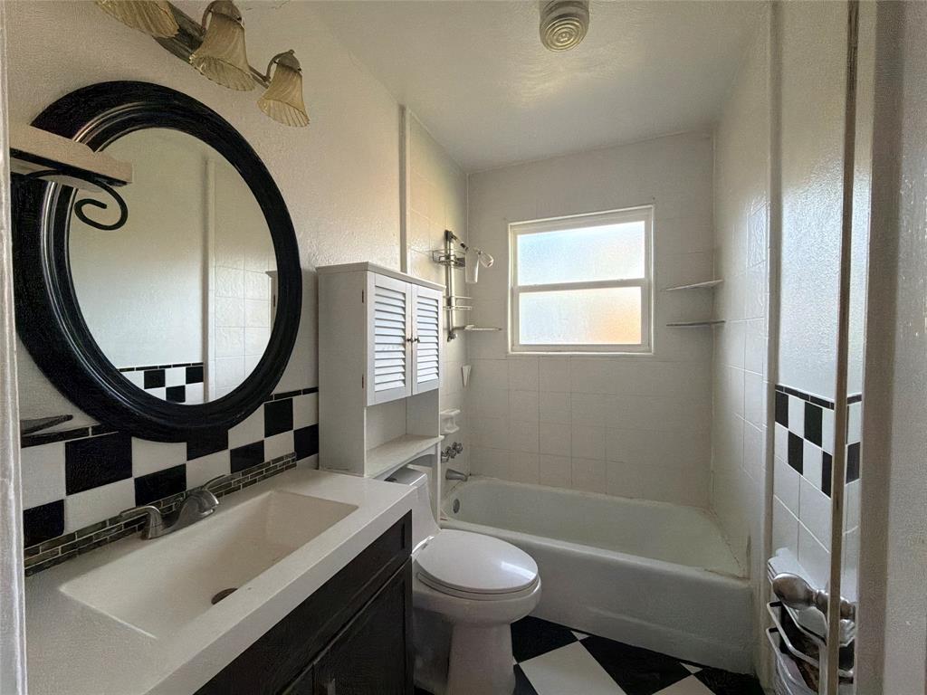 Bathroom featuring a vanity with an integrated basin, a black-framed circular mirror, a built-in shuttered cabinet, a bathtub with a tiled surround, and checkered flooring