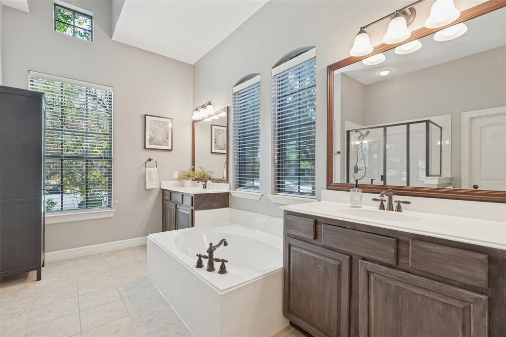Dual-sink vanity with wood-finish cabinetry, arched windows, built-in soaking tub, framed mirror, and vaulted ceiling